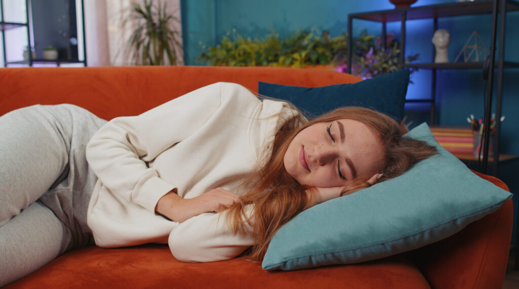 Tired caucasian adult girl lying down in bed taking a rest at home. Carefree young woman napping, falling asleep on comfortable orange sofa with pillows. Closed her eyes enjoy daytime nap alone