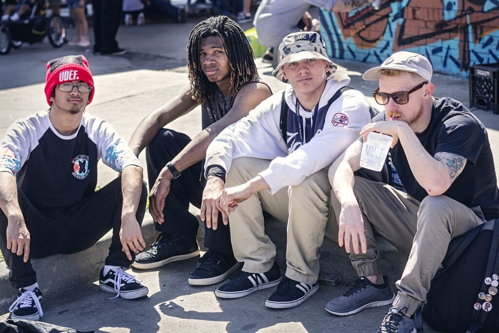 Four young men sat on the edge of a pavement One with a beanie, one with dreads, one with bucket hat an d one with baseball cap and sunglasses sipping through a straw.