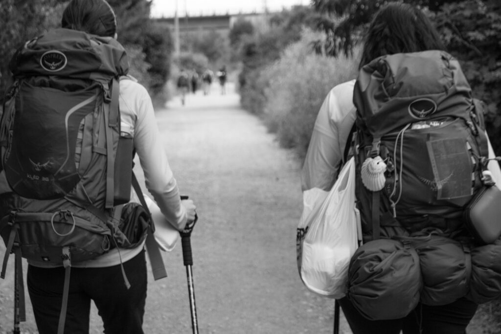 Two people walking along a path with walking poles taken from behind. Both are wearing large camping backpacks with things hanging off.