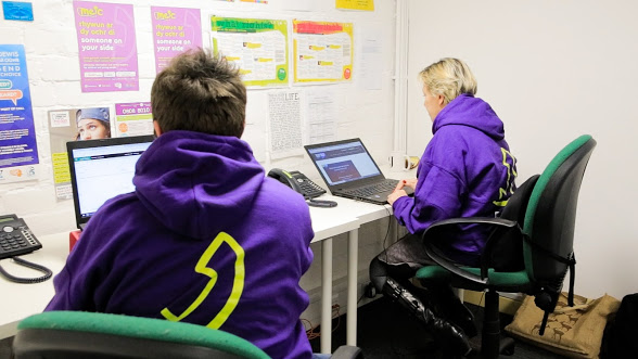 Two Meic advisers sat at their desk on laptops wearing purple Meic hoodies.