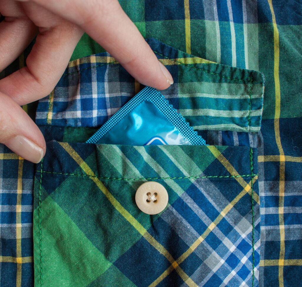 fingers pushing a condom into the breast pocket of a checked shirt, the corner of the condom is poking out
