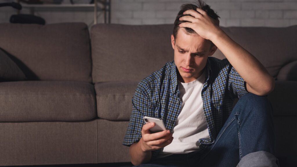 Boy sat on the floor leaning against the sofa. He has his head in his hands and is looking down at his mobile phone.