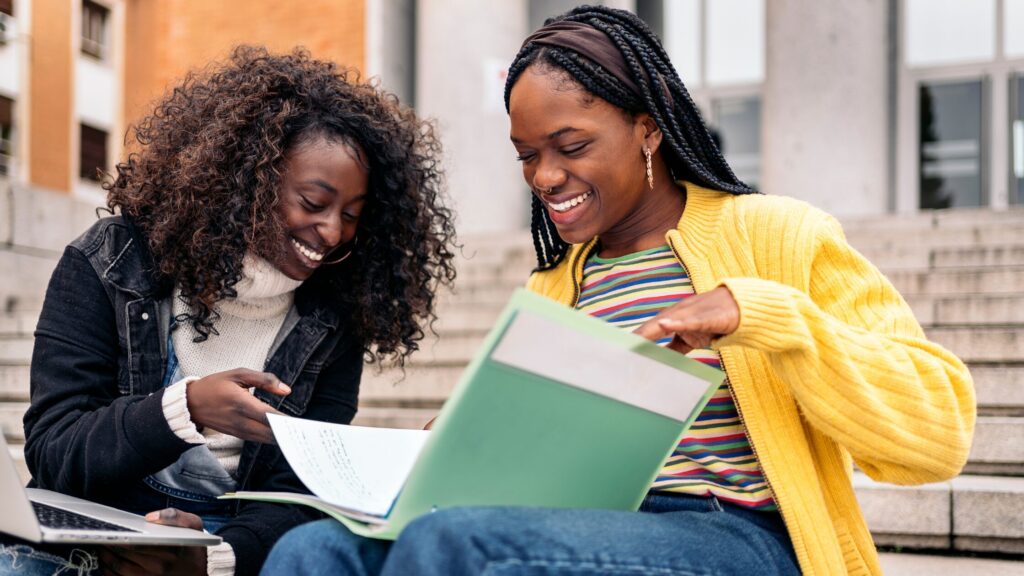 Two girls sat outdoors on stone steps. They are smiling and looking down at a shared booklet.