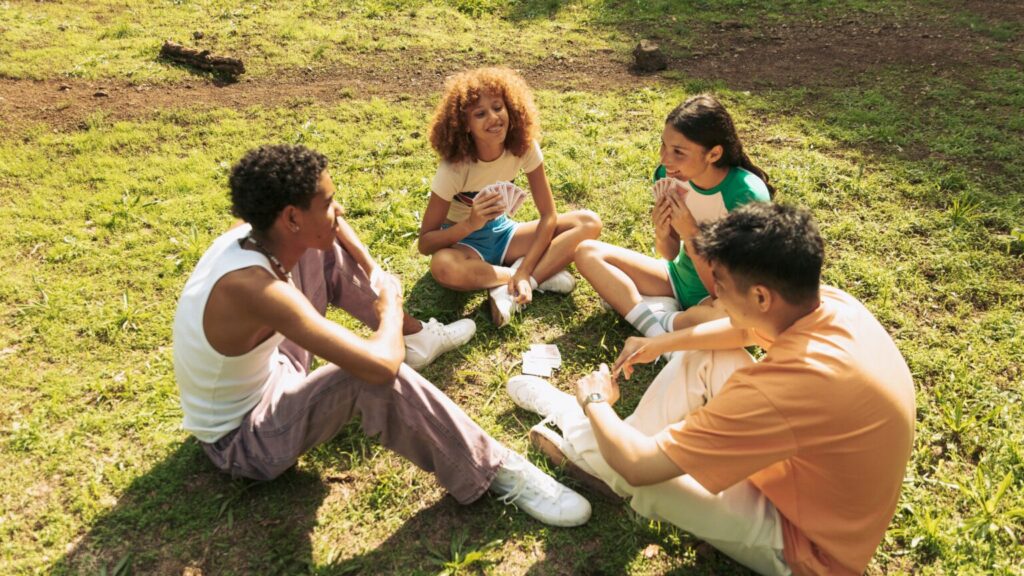 4 young people sat on the grass in a park on a sunny day playing a game of cards. There is no alcohol.