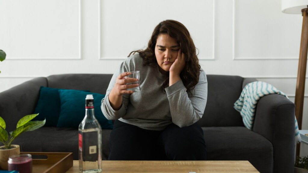 Lady sat alone on the sofa with her head in her hands. She looks at a glass filled with alcohol. A bottle of vodka is on the table in front of her.