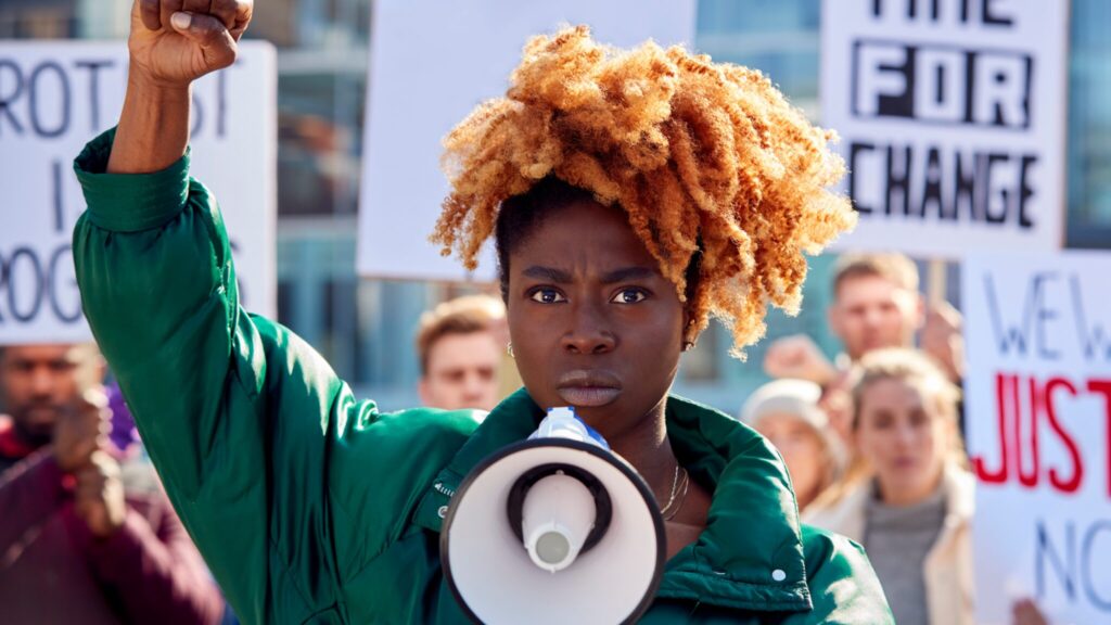 Black young adult protesting with a megaphone, with a fist raise in the air