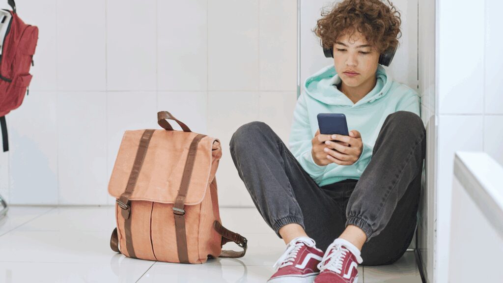 Teenager sat on the floor against a wall with mobile phone on hand, looking at websites