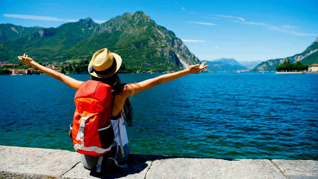 Woman wearing a sunhat and a large hiking backpack. She is sat on a stone wall, looking out over a large body of water and a luscious, rocky and green mountain.