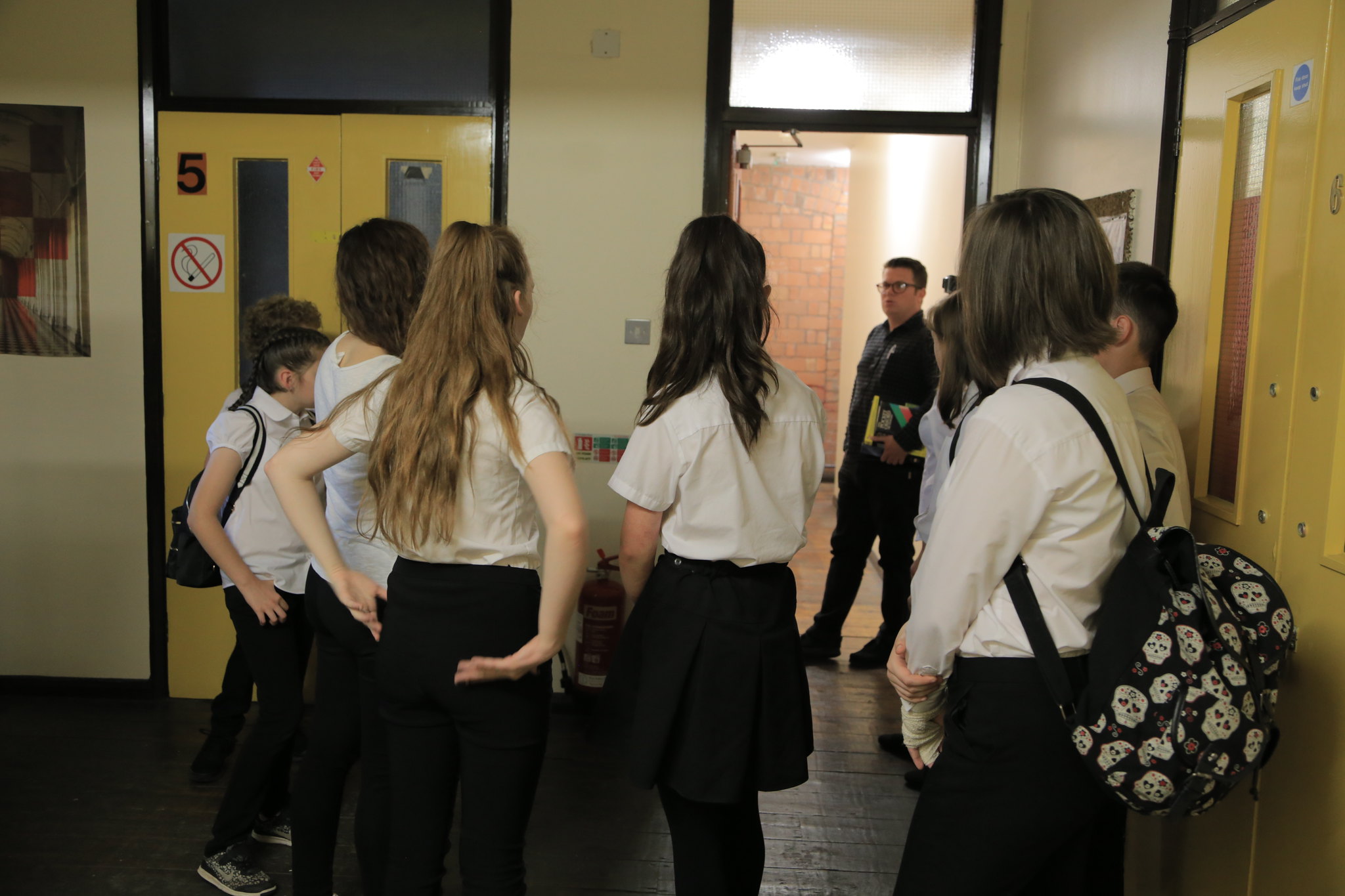 A group of young people in school uniform stood outside some rooms in a building with a man stood in front of them with books under his arm.