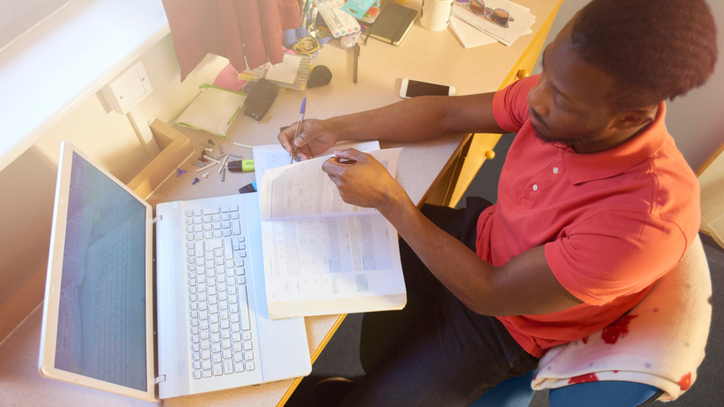 Young man studying at a cluttered desk with a laptop and open textbook.