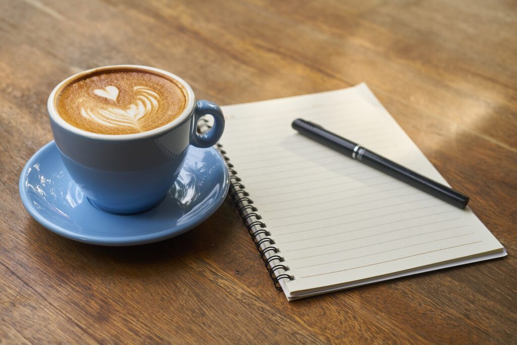 Cup of coffee with milk art on top of a heart. Lined notebook open with pen resting on top.