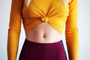 A belly button piercing on someone wearing a yellow sie front top and burgundy bottoms. Only their torso is visible