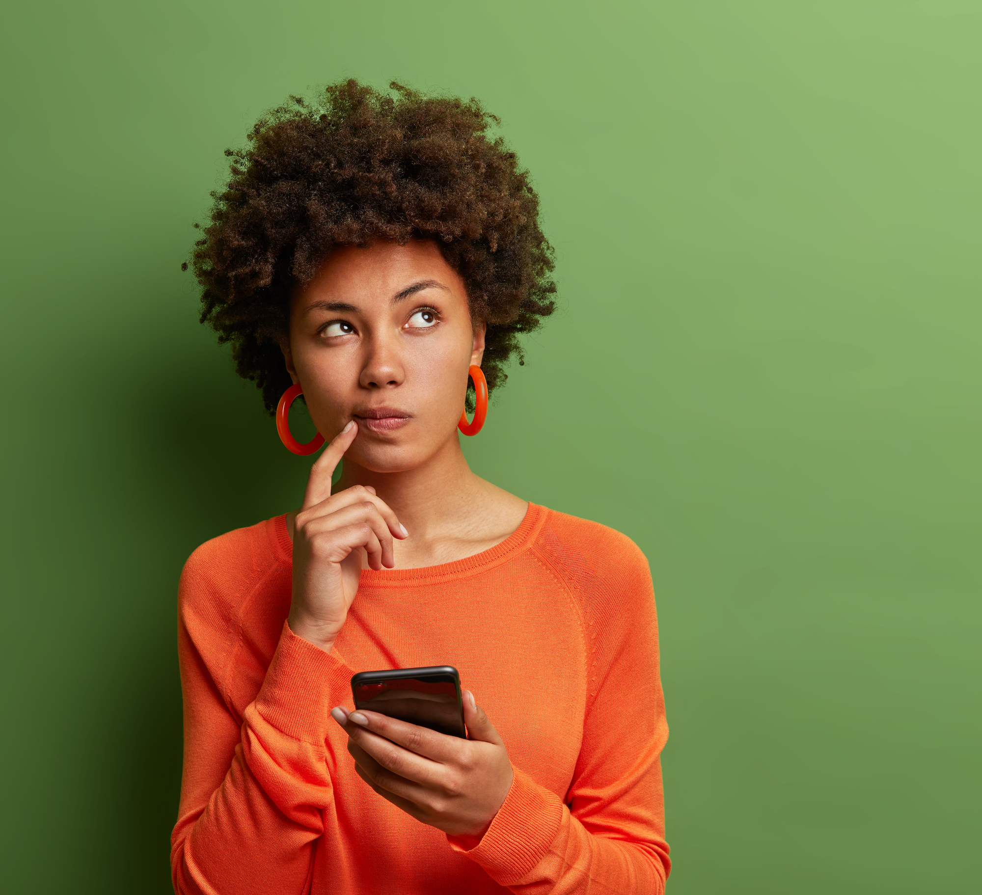 Photo of pretty ethnic woman ponders on how to answer question, thinks deeply about something, uses modern mobile phone