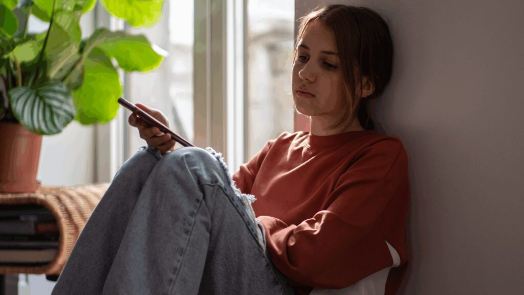 Teen girl sat slouched against a wall with phone in hand, looking sad