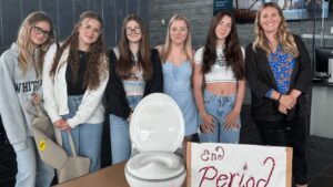 A group of six females, stand behind a display raising awareness for period dignity. The display features a white portable commode and a hand-drawn sign with red lettering that says "End Period."
